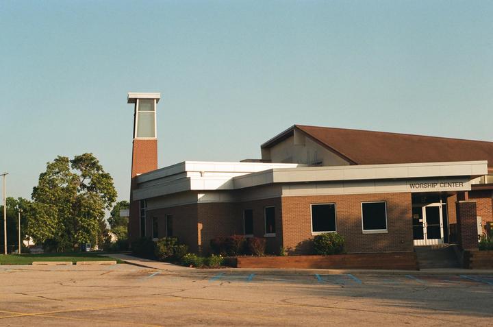 A brick building with a tall brick tower and a sign that reads "WORSHIP CENTER" stands next to a tree and a parking lot.