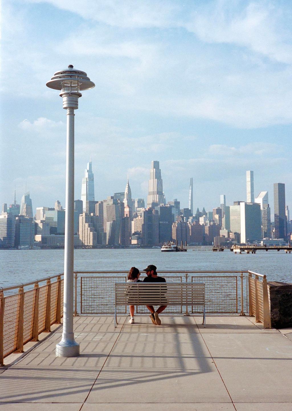 A couple sits on a bench by the water with a city skyline in the background.