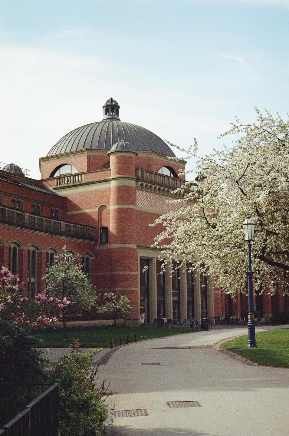 A large brick building with a domed roof stands beside a winding path and flowering trees.