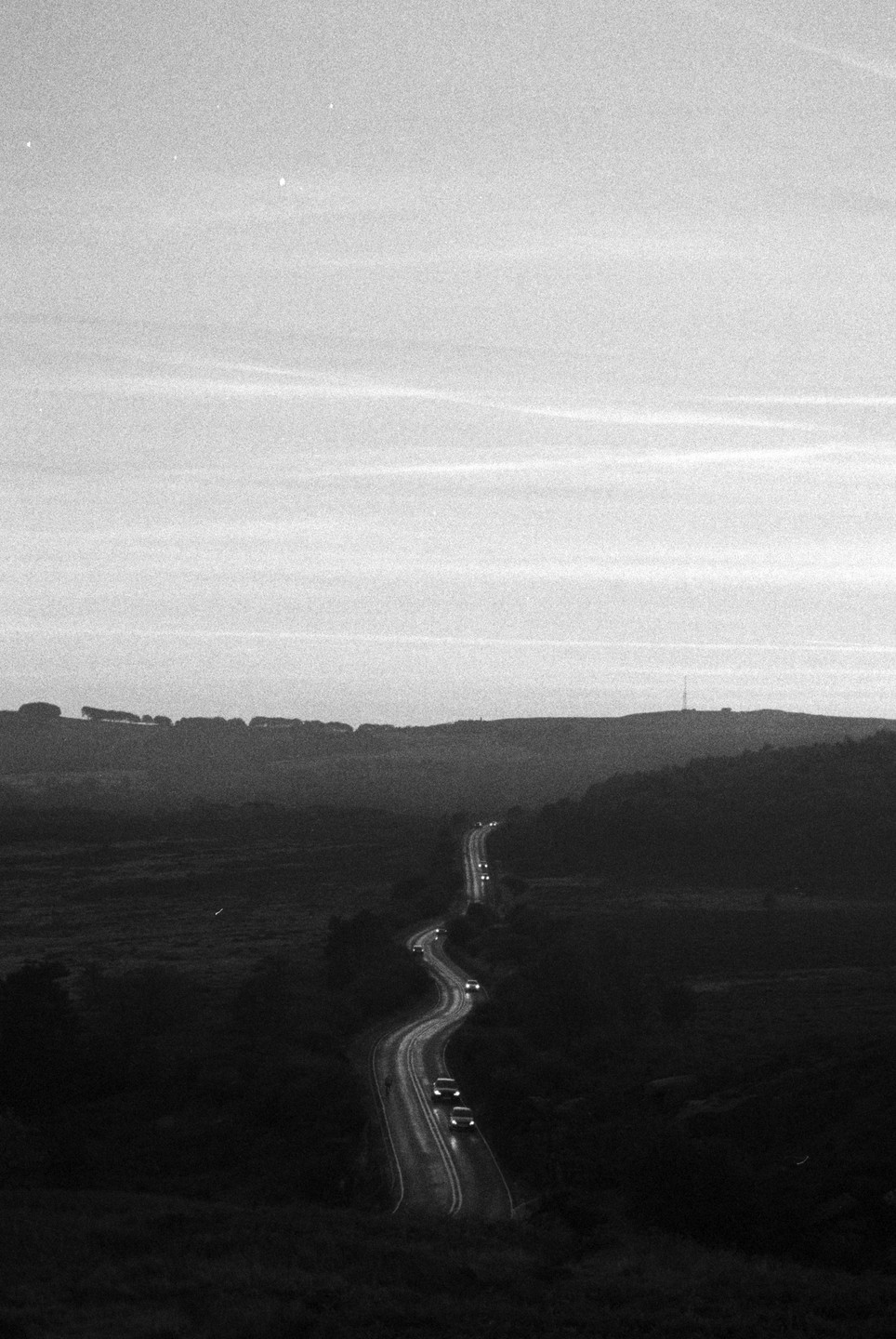 A winding road with cars driving on it cuts through a dark, hilly landscape under a bright sky.