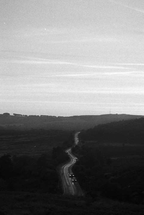 A winding road with cars driving on it cuts through a dark, hilly landscape under a bright sky.