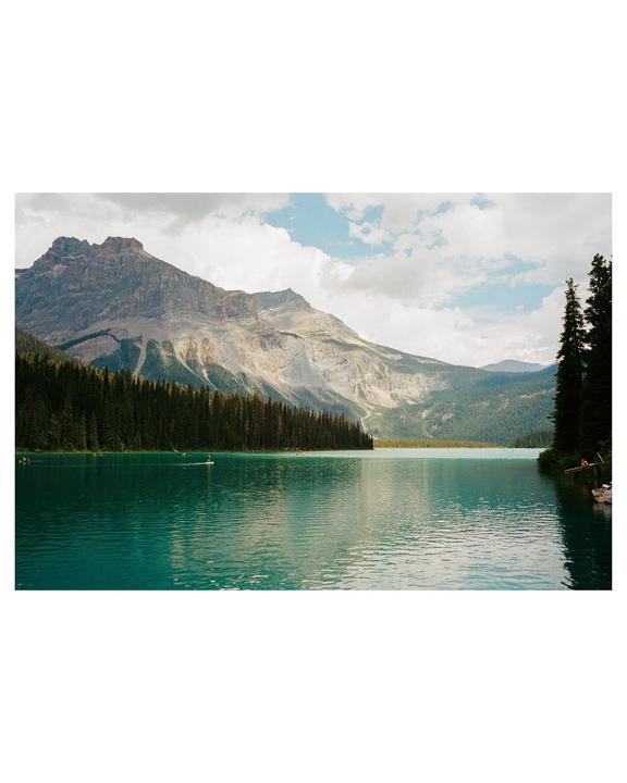A turquoise lake is surrounded by a dense forest and a large mountain under a cloudy sky.