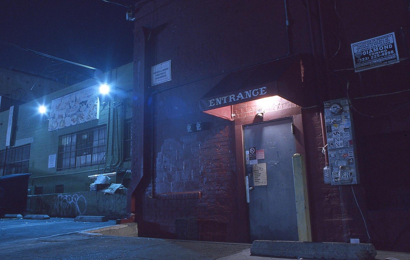 An alleyway at night shows a building with an "ENTRANCE" sign above a door and another building with a mural and barred windows.