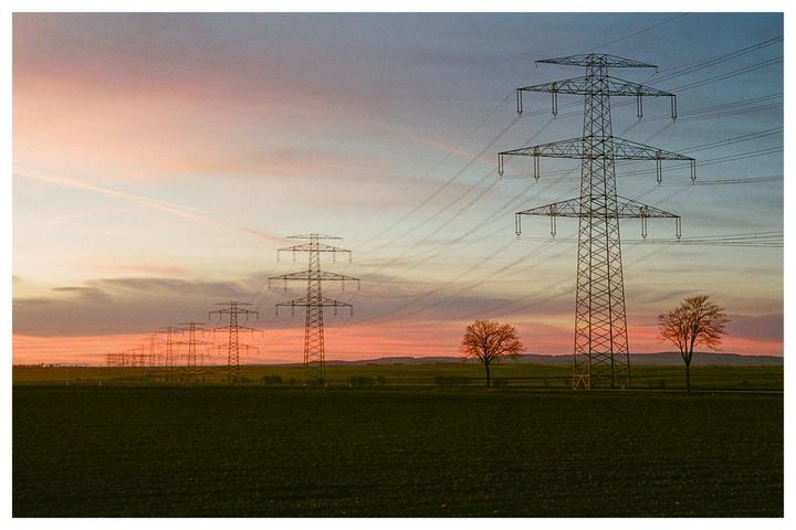 A row of electricity pylons stretches across a field under a colorful sunset sky.