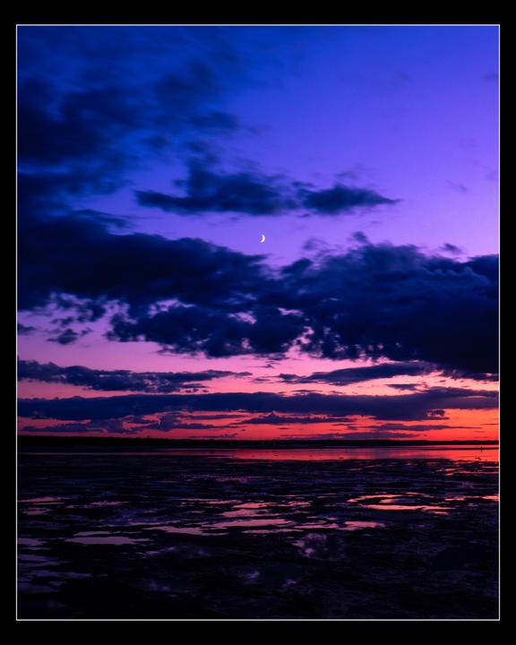 A crescent moon is visible in the purple and red sky over a body of water at sunset.