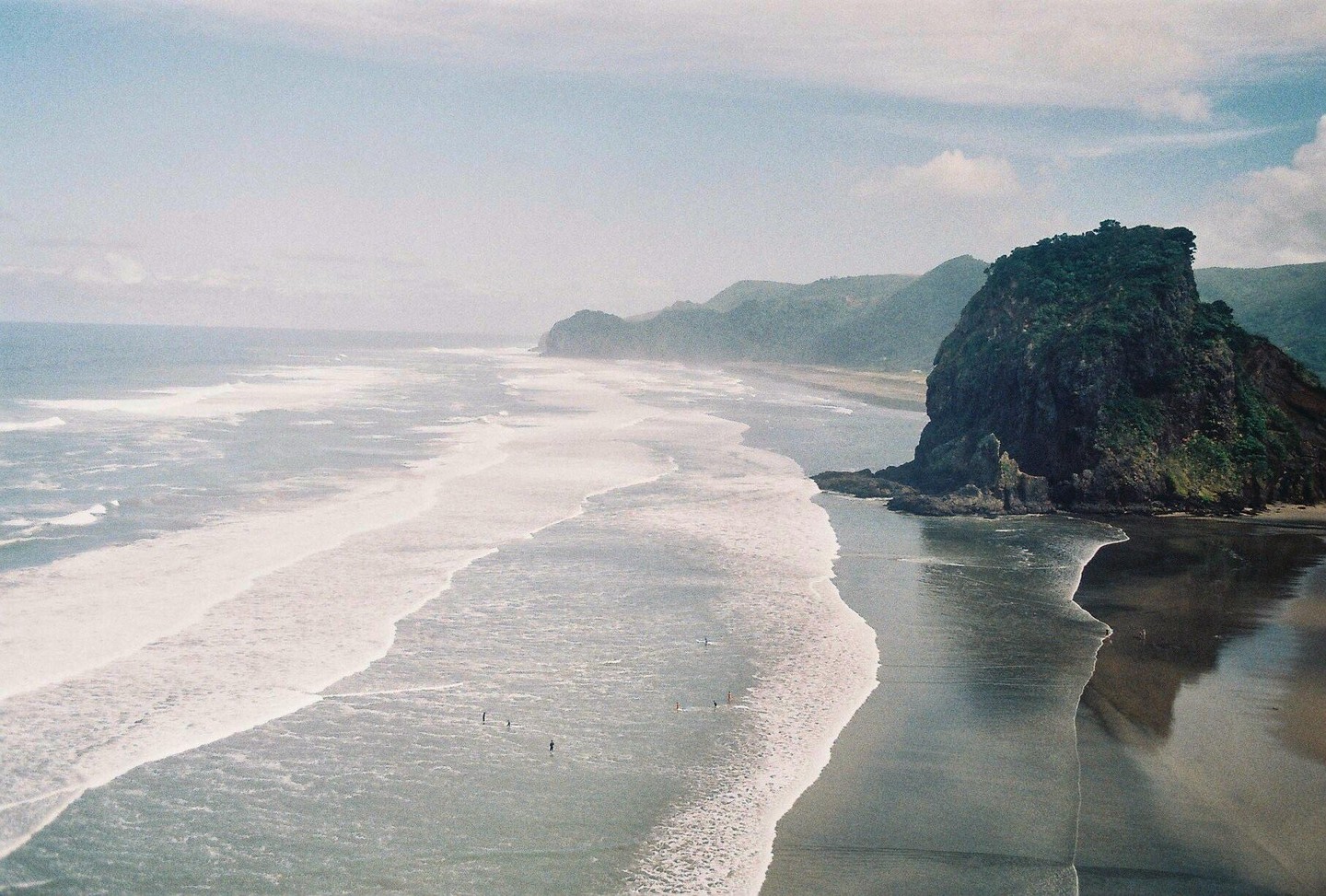 A wide shot of a beach with waves rolling in, a large rock formation on the right, and mountains in the distance.