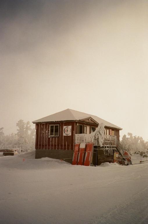 A red wooden cabin is covered in snow with snow-covered trees in the background.