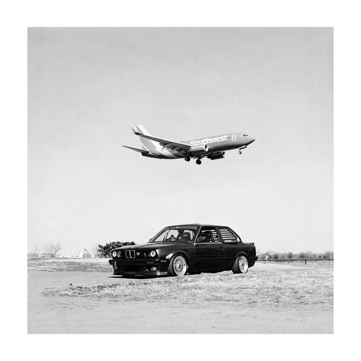 A black car is parked on a field with an airplane flying overhead.