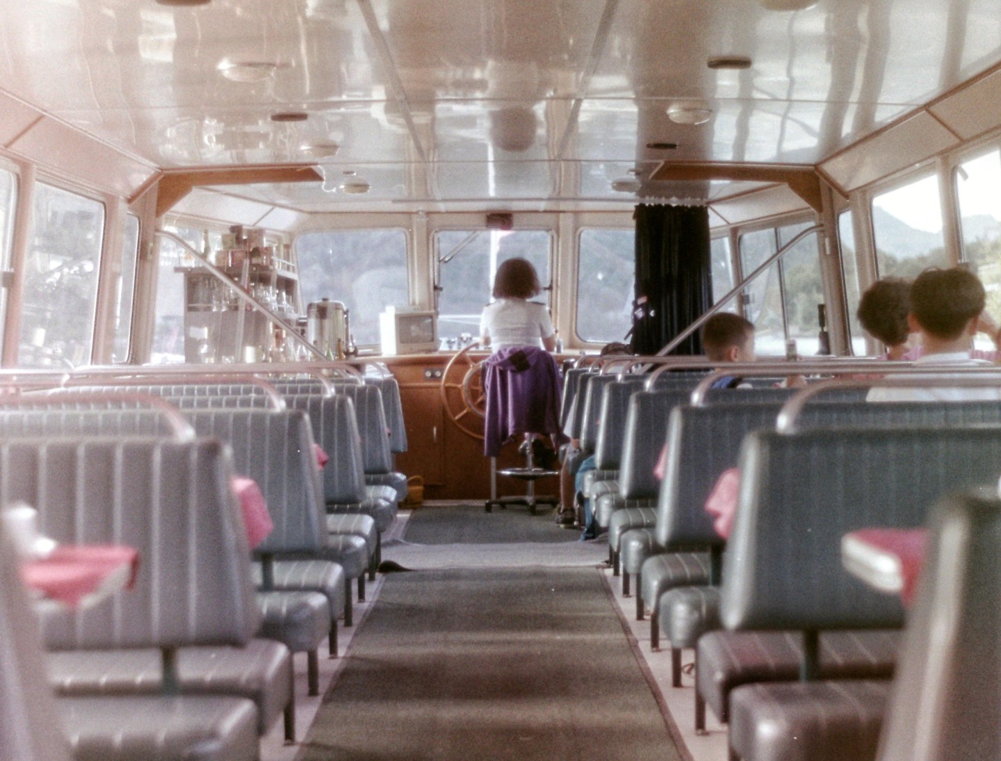 A woman stands at the helm of a ferry with passengers seated in rows behind her.