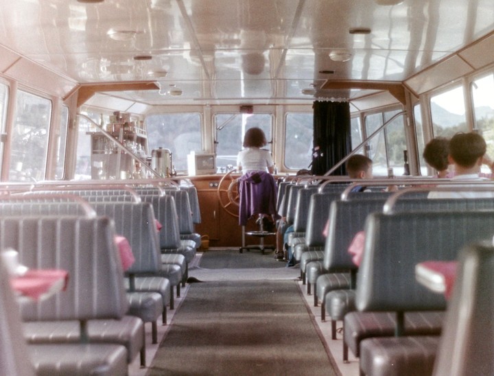 A woman stands at the helm of a ferry with passengers seated in rows behind her.