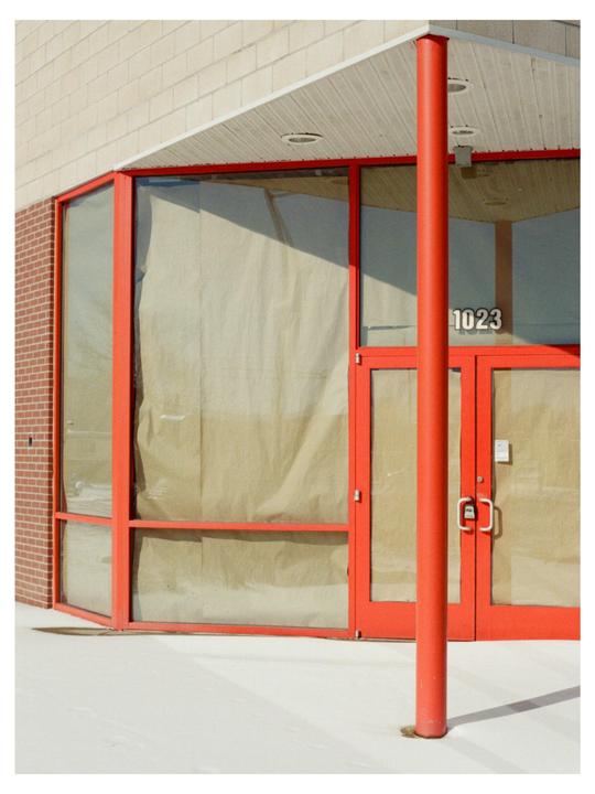 A red pillar stands in front of a building with red-framed windows and a door, with the number 1023 visible on the door.
