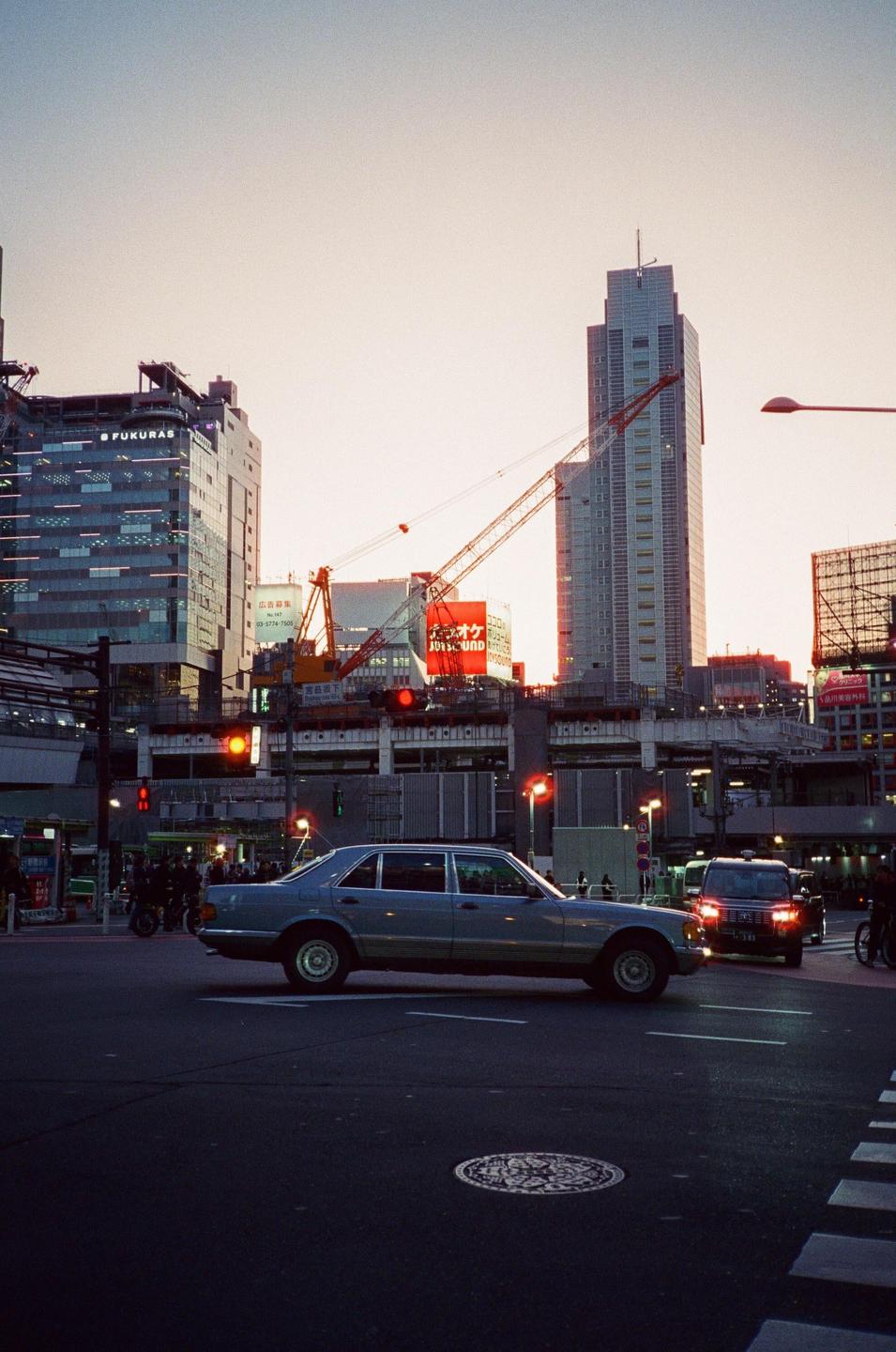 A car is stopped at an intersection in a city with tall buildings and construction cranes in the background.