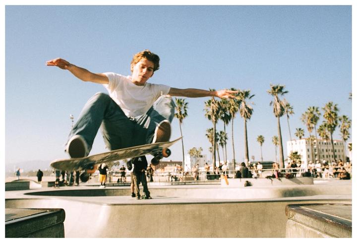A young man is captured mid-air on a skateboard at a skate park with palm trees and buildings in the background.