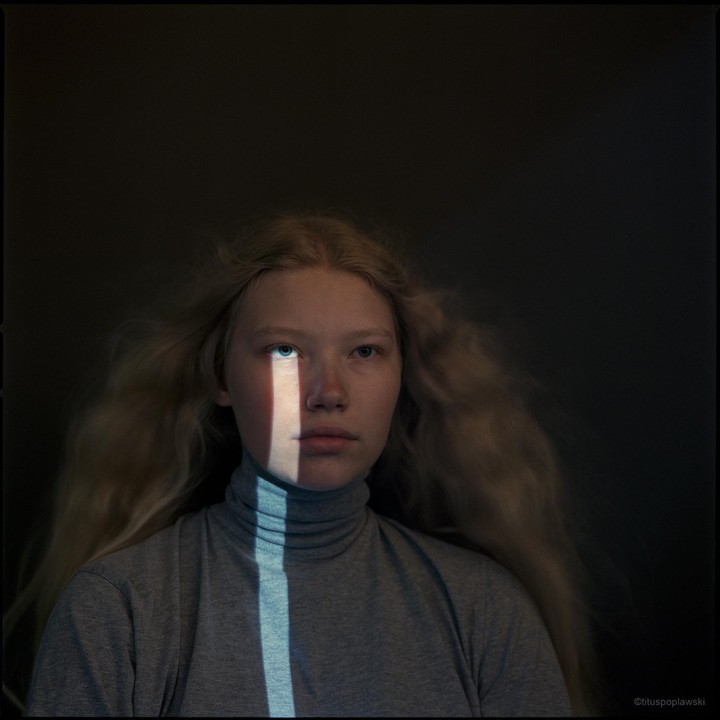 A young woman with long blonde hair wears a grey turtleneck, with a vertical beam of light illuminating part of her face and neck.