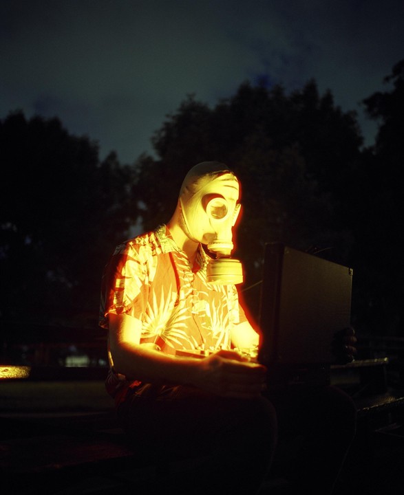A person wearing a gas mask and a Hawaiian shirt sits in the dark, illuminated by a glowing briefcase.