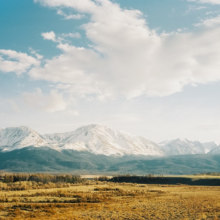 Snow-capped mountains rise above a valley with trees and fields under a cloudy sky.