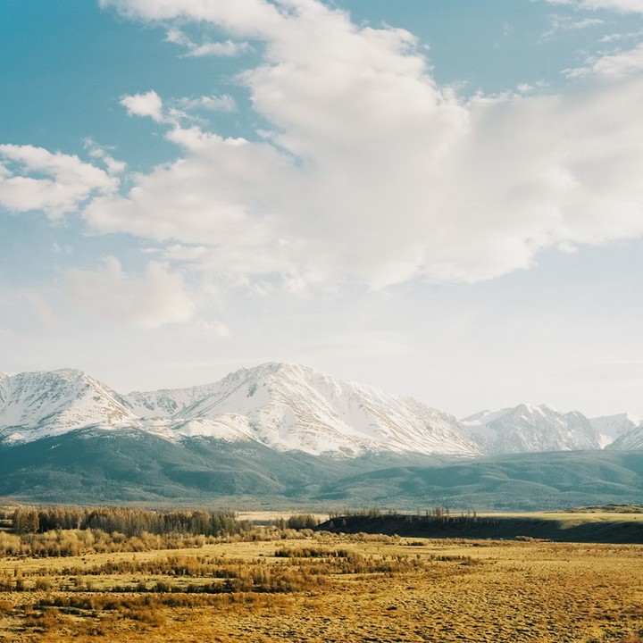 Snow-capped mountains rise above a valley with trees and fields under a cloudy sky.