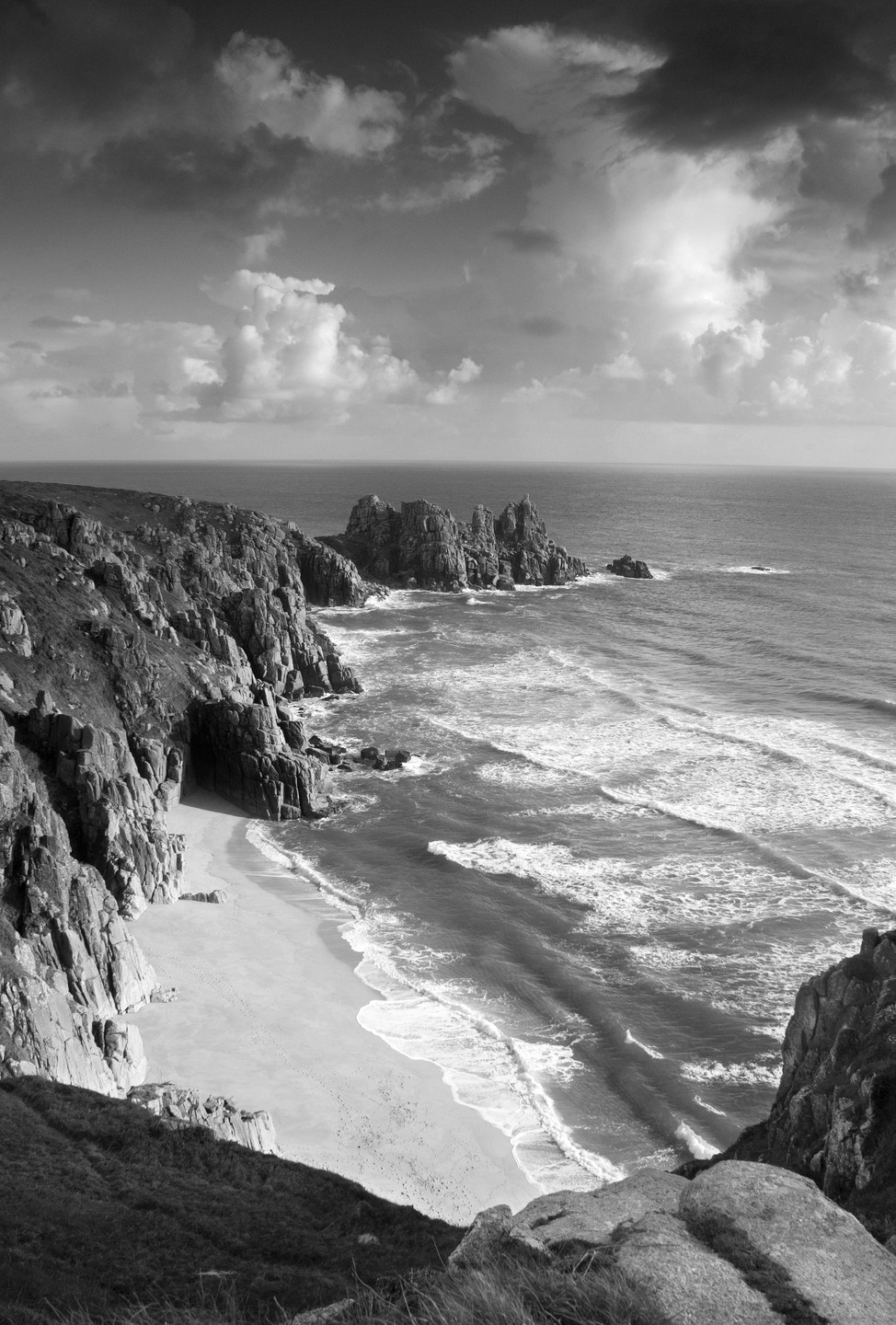 A black and white landscape shows a sandy beach at the base of a rocky cliff with waves breaking on the shore and a cloudy sky overhead.