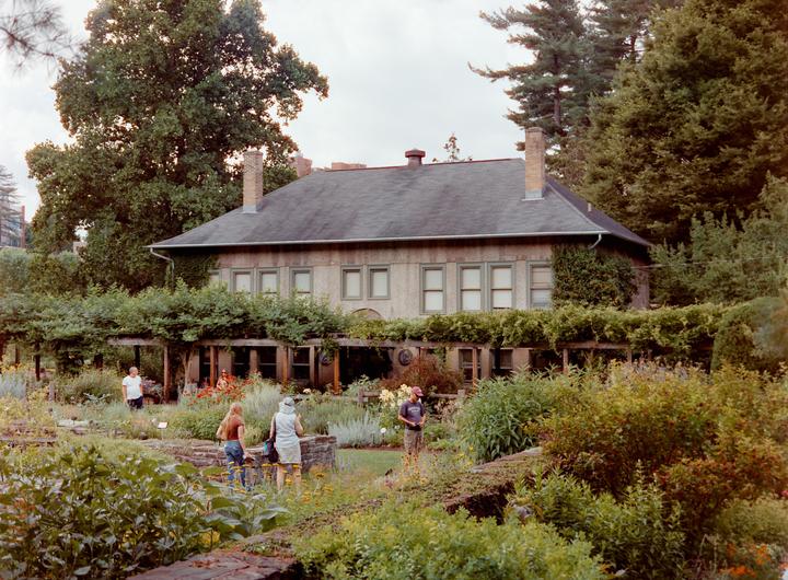 A large house with a dark roof and many windows is surrounded by lush green gardens and trees, with several people walking through the garden.
