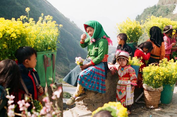 A woman and several children are gathered outdoors among baskets of yellow flowers with a mountain in the background.