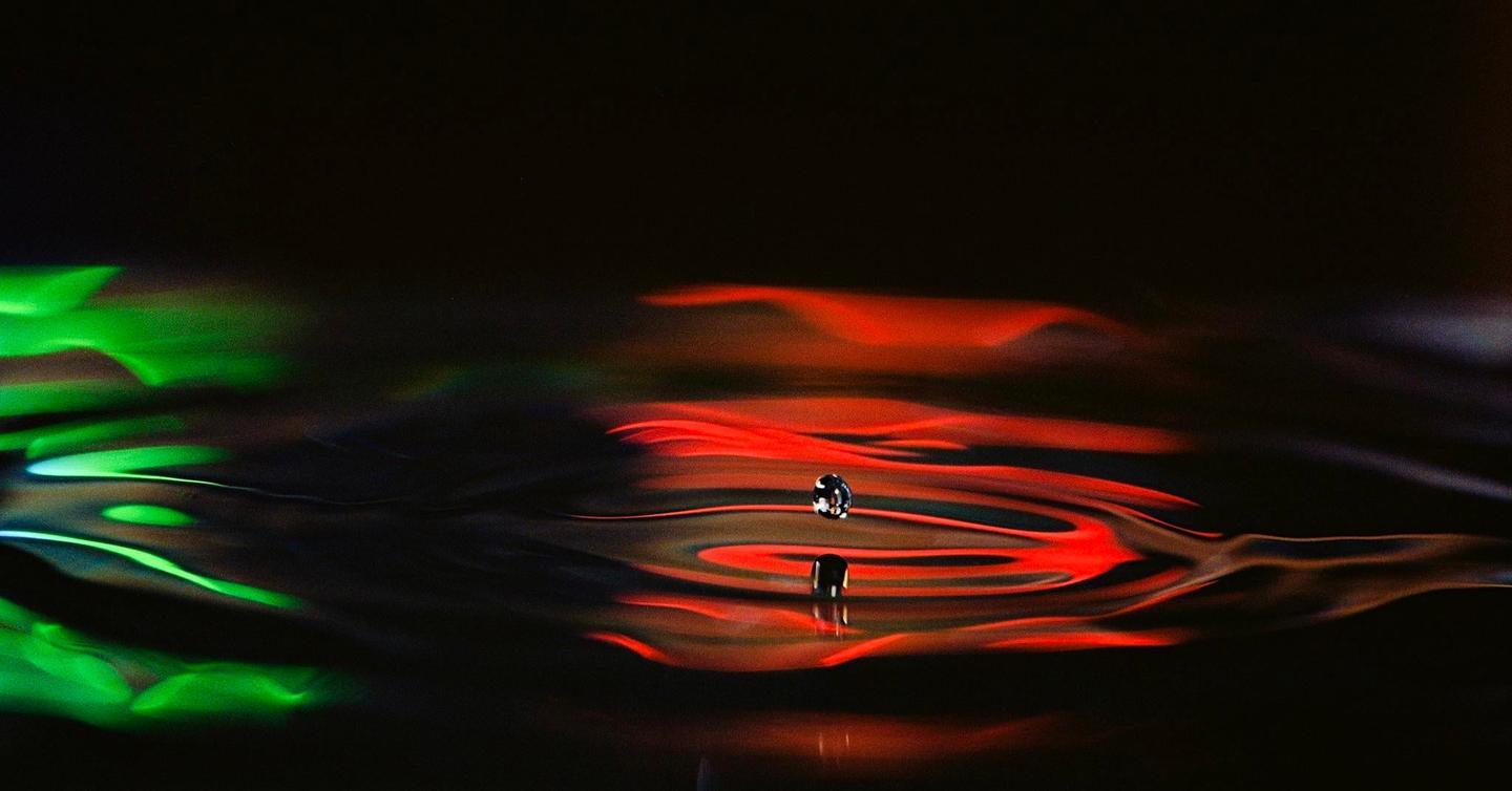 A water drop is suspended above a surface with colorful reflections, while another drop is reflected below.