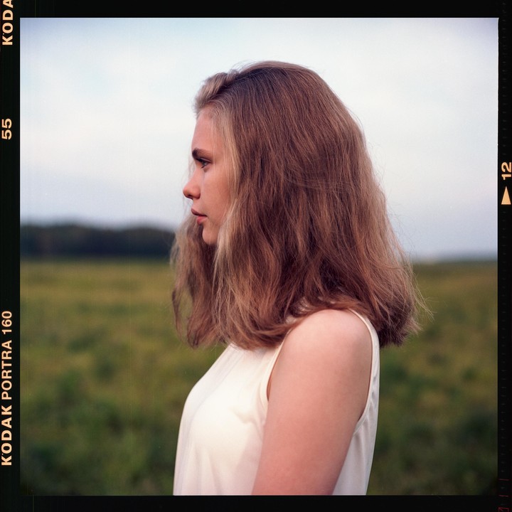 A young woman with long hair stands in profile in a field.