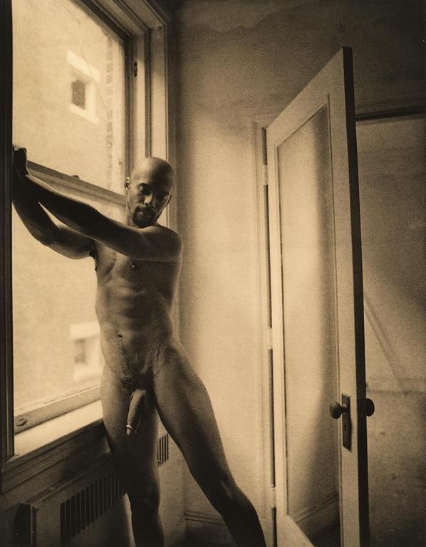 A nude man stands by a window with an open door to his right.
