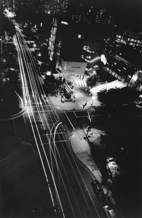 A high-angle, black and white night photograph captures a city street with light trails from moving vehicles.