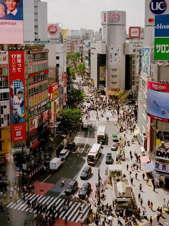 A high-angle view shows a busy street in a city with many pedestrians, vehicles, and buildings covered in advertisements.