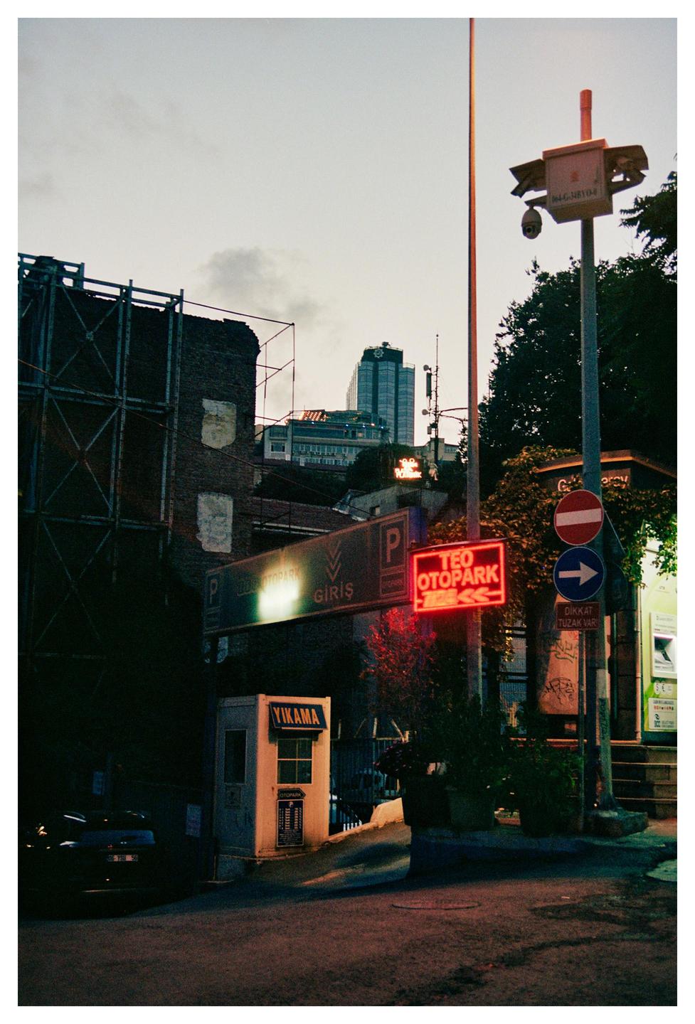 A street scene at dusk shows a parking garage entrance with neon signs and a tall building in the background.