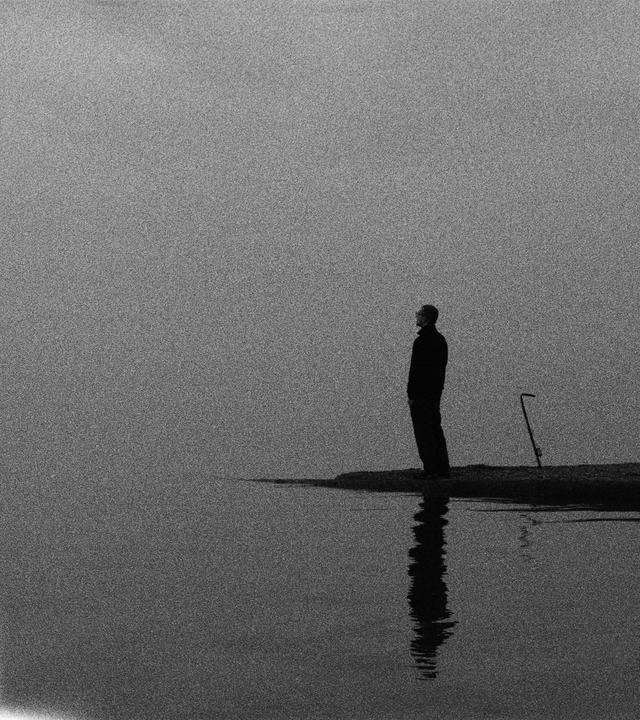 A man stands on a small strip of land next to water, with a fishing rod nearby.