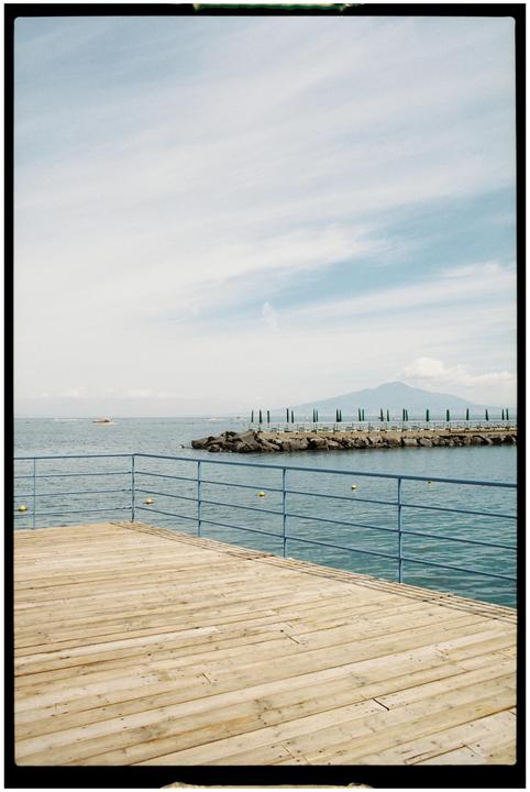 A wooden pier with a blue railing extends over the water towards a rocky breakwater lined with green beach umbrellas and a distant mountain under a cloudy sky.