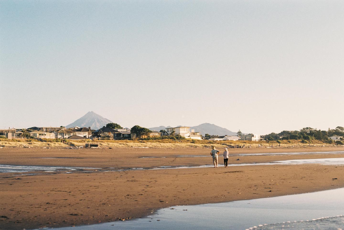 Two people walk on a wide sandy beach with houses and a mountain in the distance under a clear sky.