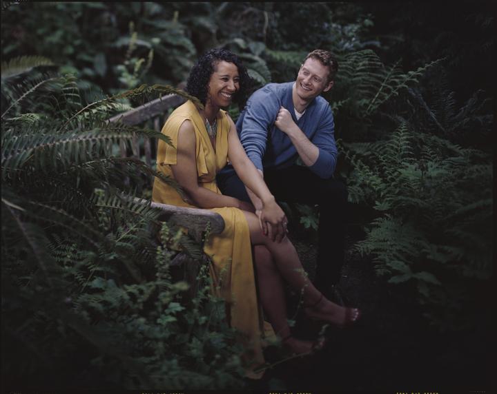 A couple sits on a wooden bench surrounded by ferns.