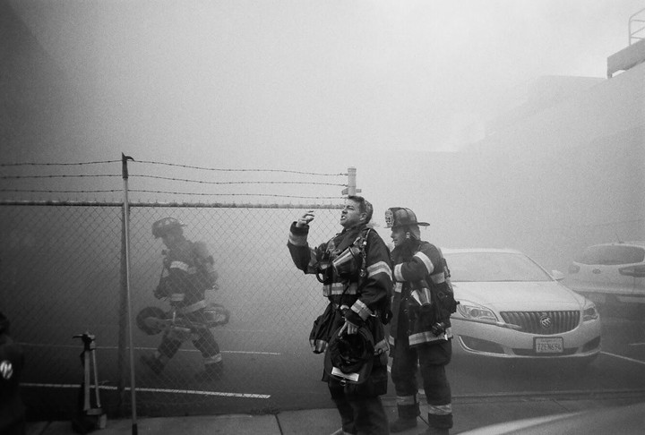 Three firefighters stand in a hazy street next to a chain-link fence and a car.