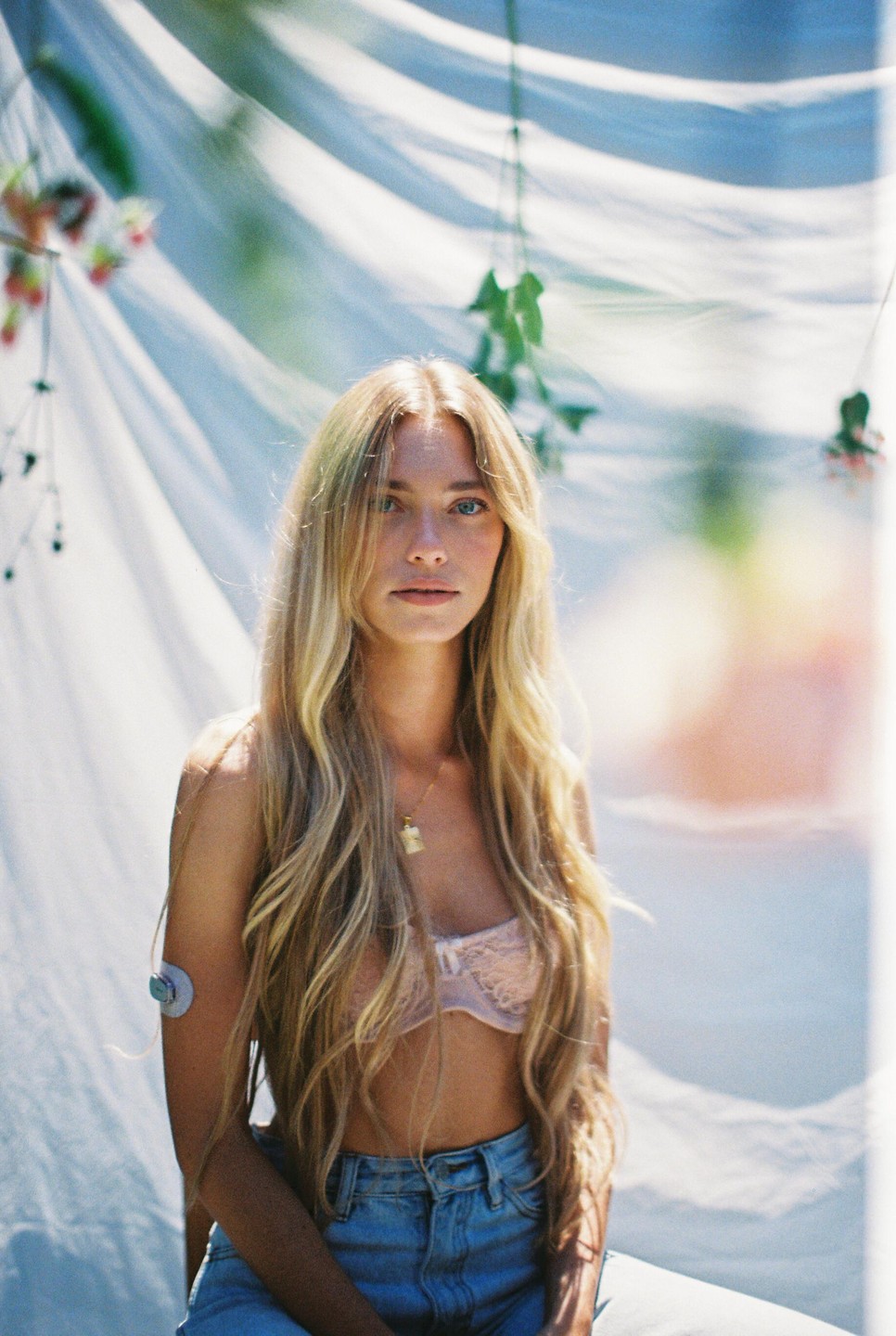 A woman with long blonde hair and a pink bra sits in front of a white sheet with green vines.