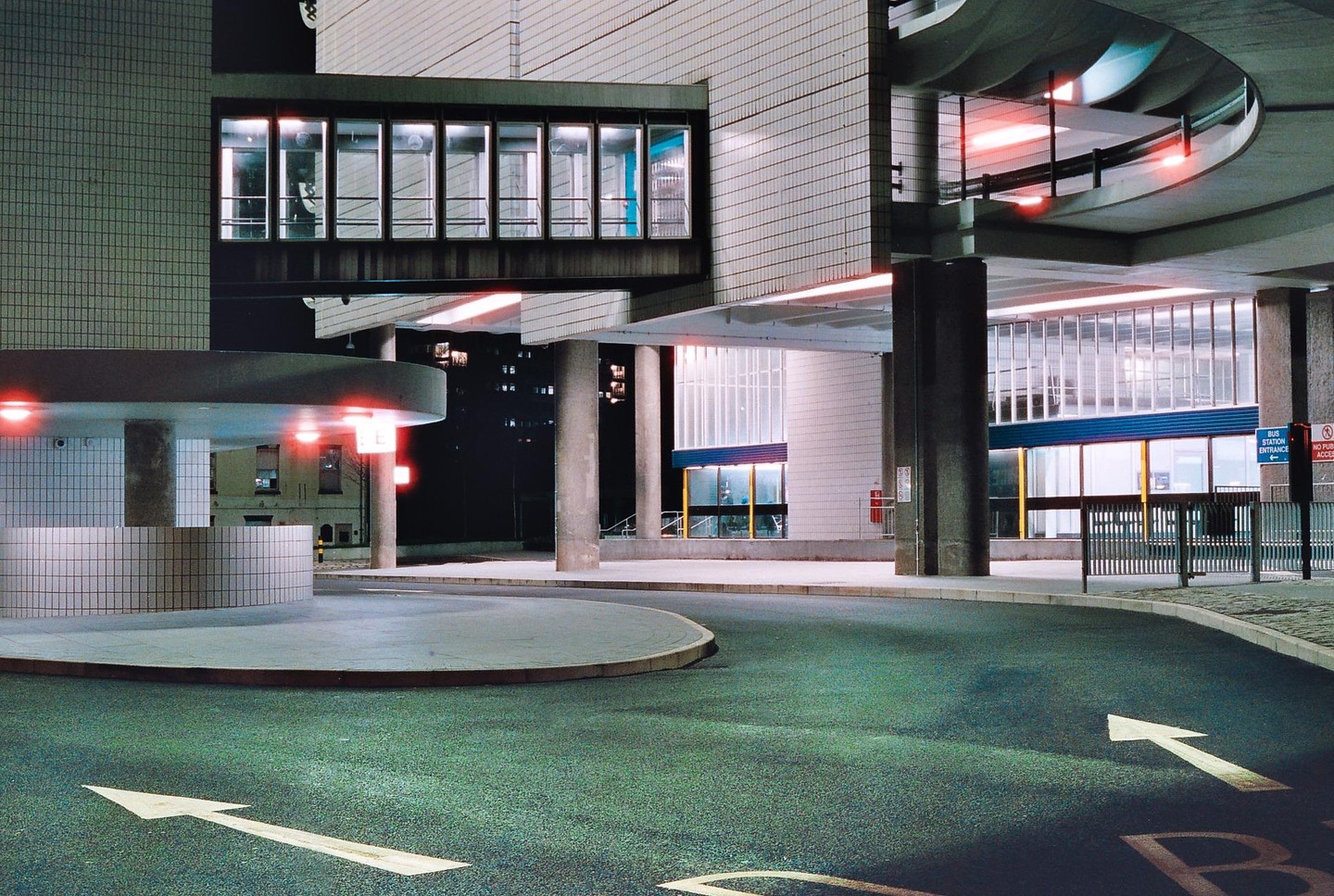 A modern building with a skybridge and a circular drive with arrows painted on the asphalt is illuminated at night.
