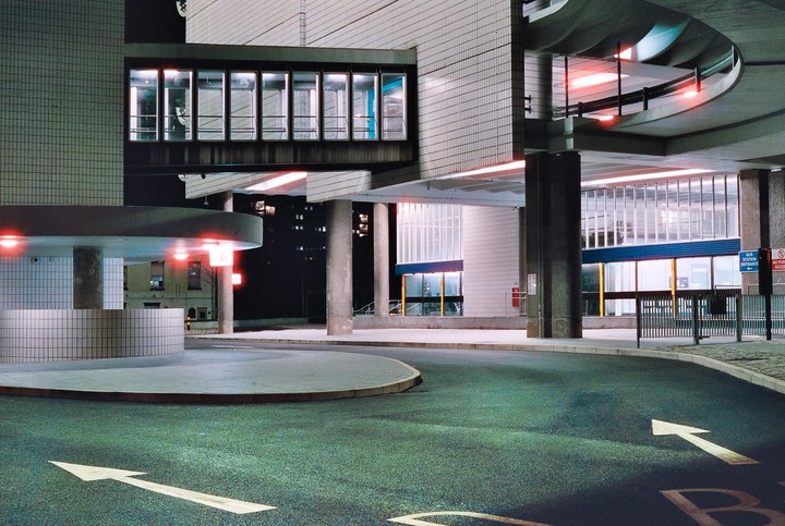 A modern building with a skybridge and a circular drive with arrows painted on the asphalt is illuminated at night.