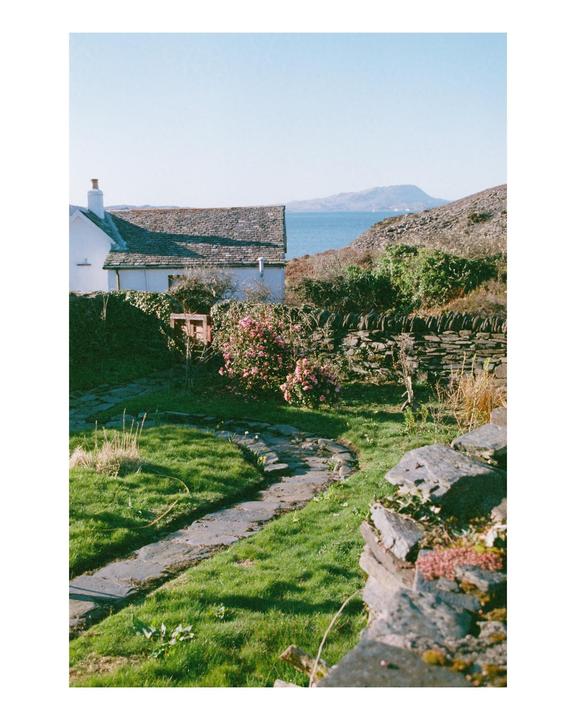 A stone path winds through a grassy garden towards a white house with a view of the sea and mountains in the distance.
