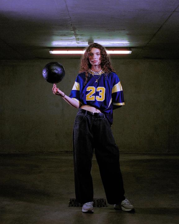 A woman in a blue and yellow jersey balances a black soccer ball on her finger in a dimly lit room.