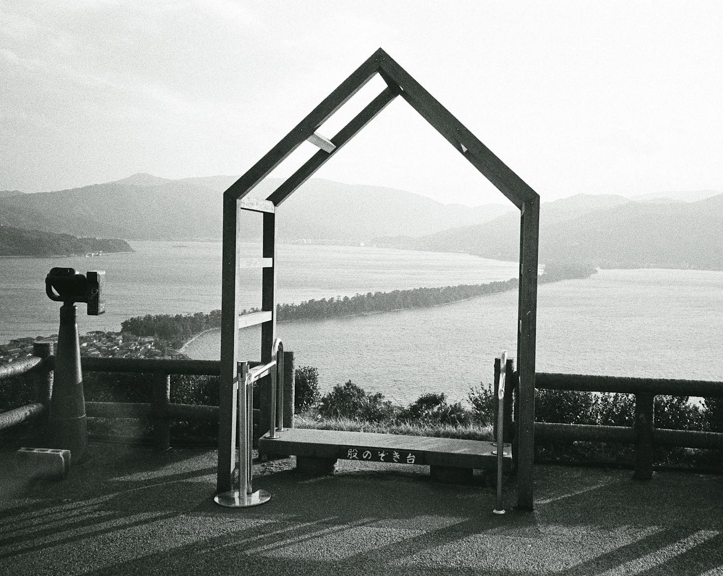 A wooden frame shaped like a house stands on a hill overlooking a body of water with a narrow land bridge and mountains in the distance.