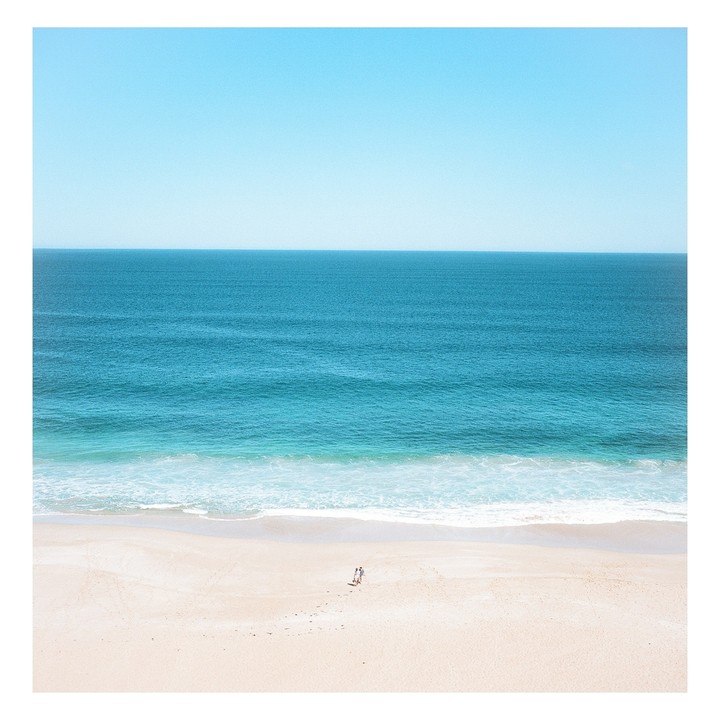 A couple walks on a vast, empty beach next to the ocean under a clear sky.