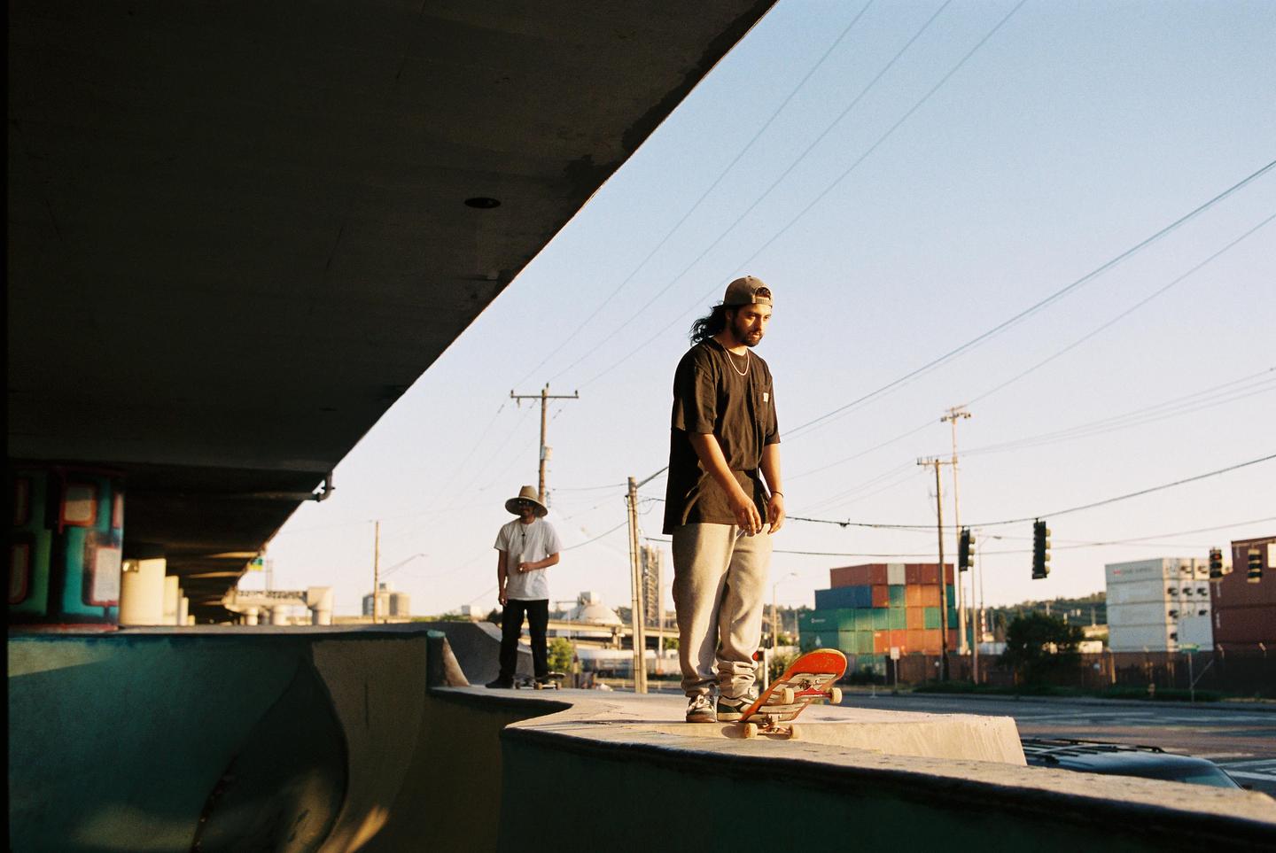 Two men stand with skateboards in a skatepark under a bridge with shipping containers in the background.