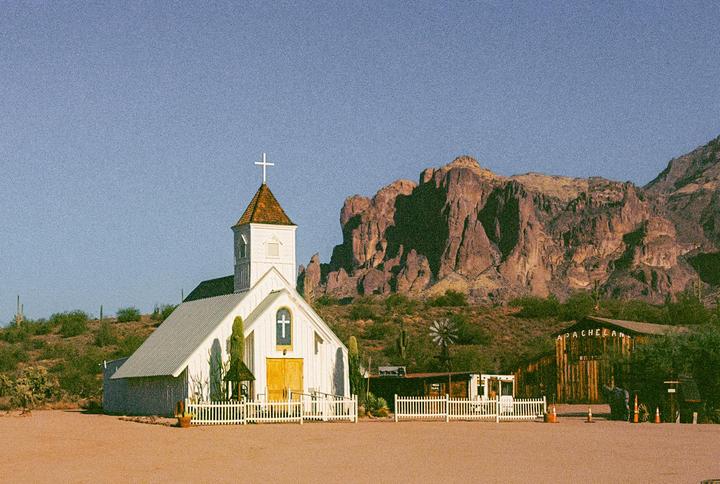 A white church with a steeple stands in a desert landscape with mountains in the background.