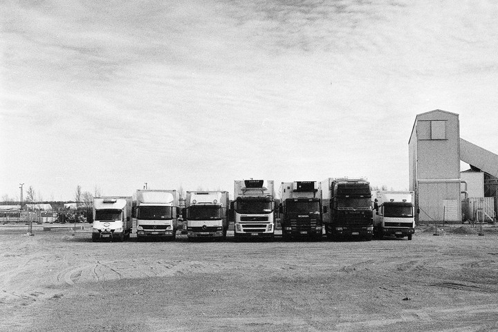 Six trucks are parked in a row on a dirt lot next to an industrial building.