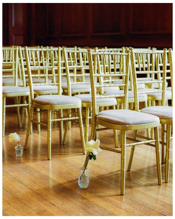 Rows of golden chairs with white roses in small vases are arranged on a wooden floor.