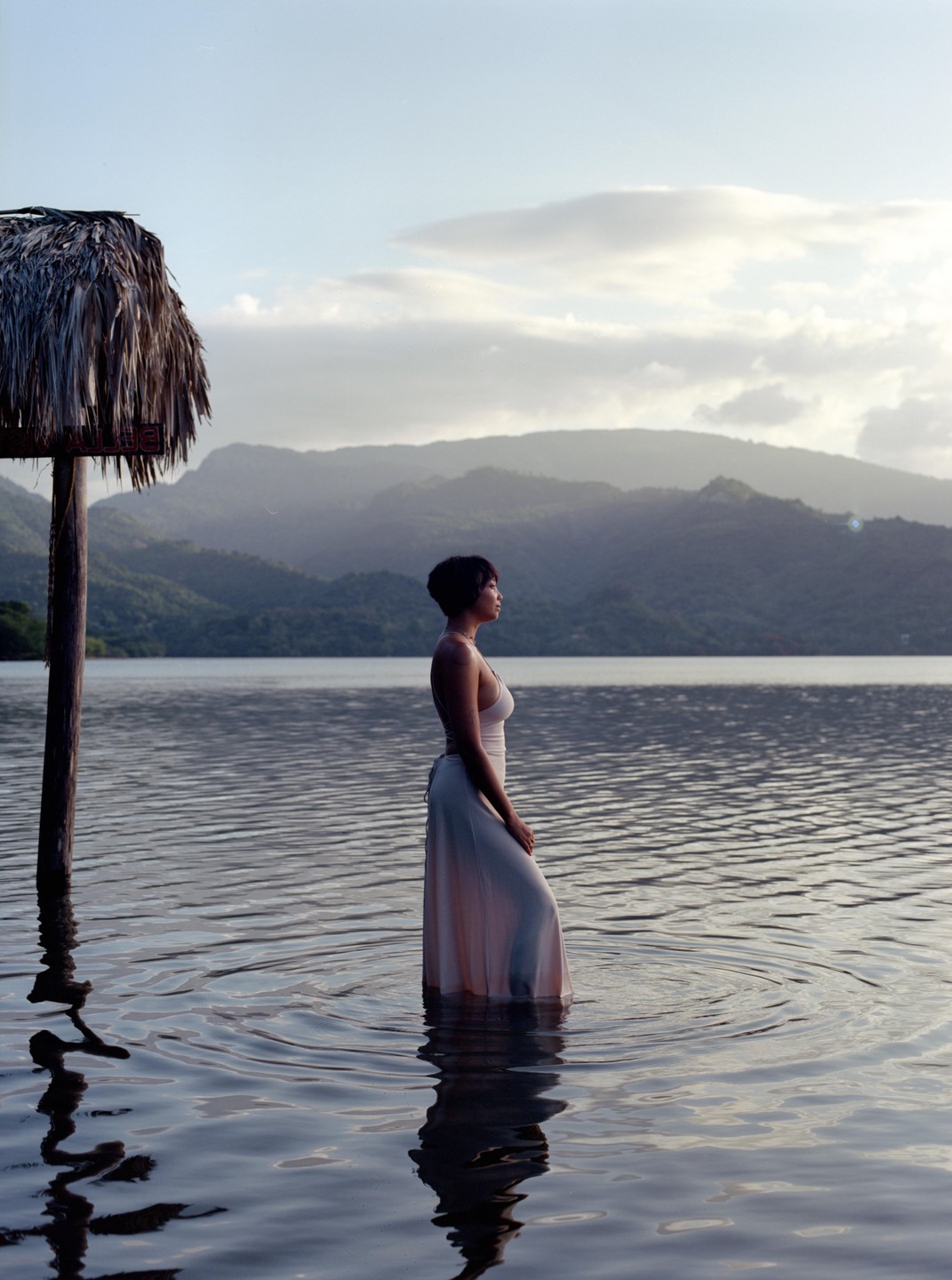 A woman stands in a body of water with mountains in the background and a thatched post to her left.
