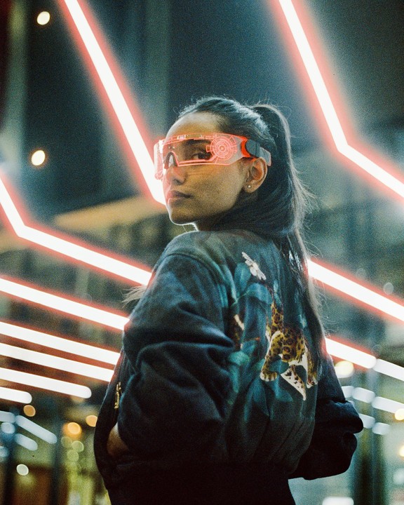 A woman wearing futuristic glasses and a jacket with an animal print stands in front of neon lights.