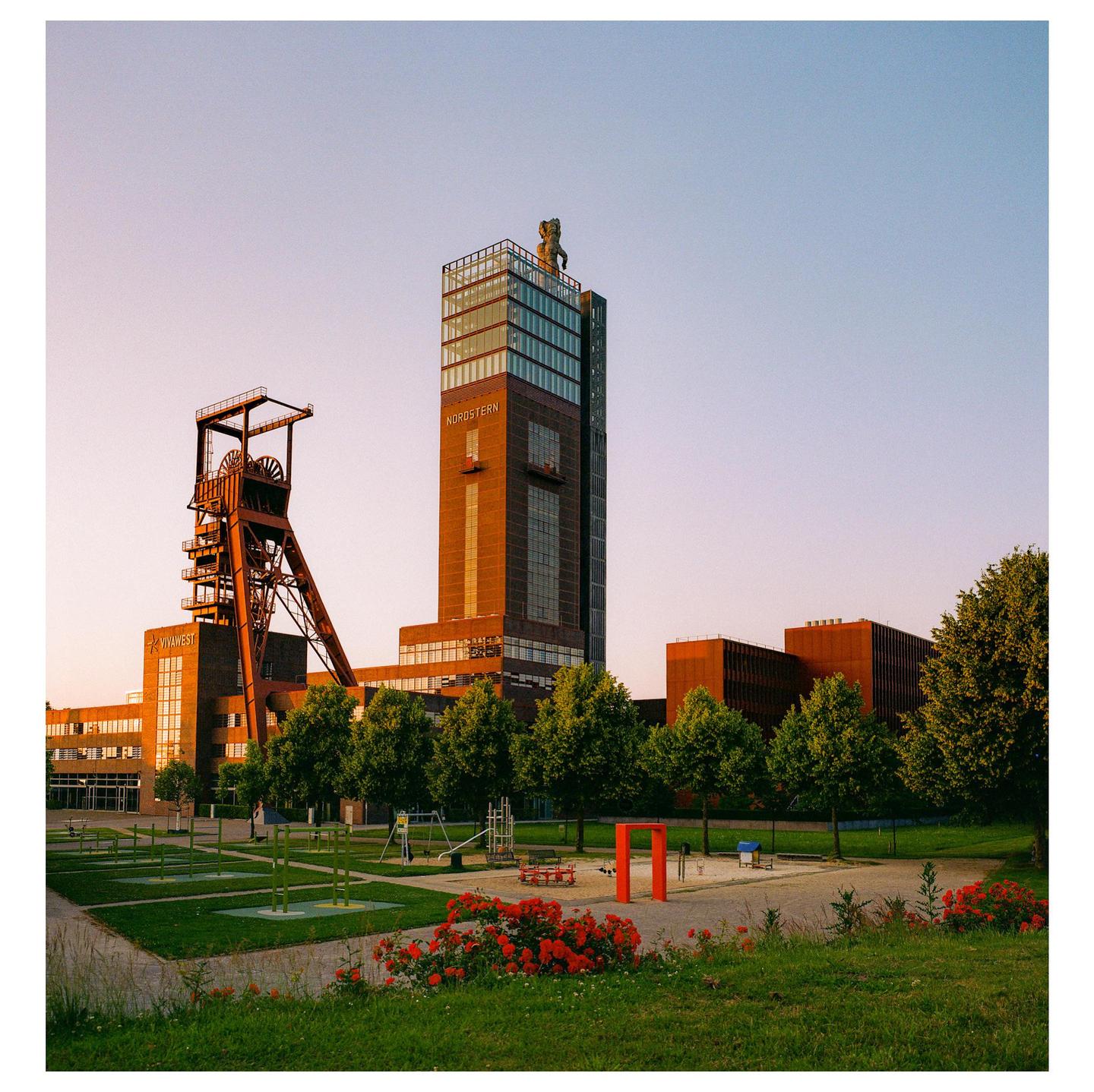 A tall building with "NORDSTERN" written on it stands next to an industrial structure, with a park and red flowers in the foreground.