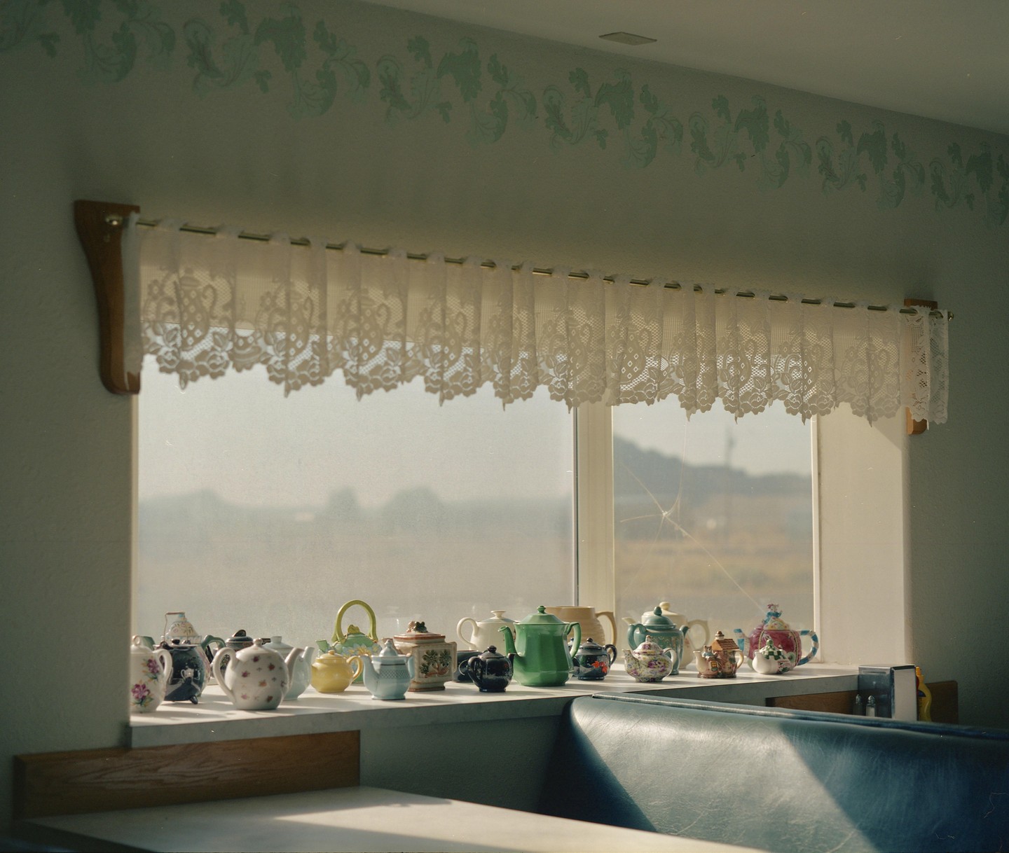 A collection of decorative teapots sits on a windowsill with a lace curtain above them.